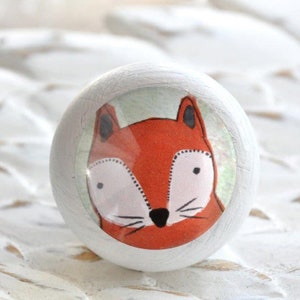 Könnte beinhalten: Weißer Holz-Knopf mit einem runden, bunten Aufkleber, der einen Comic-Fuchs mit orangefarbenem Fell, schwarzen Augen und einer rosa Nase zeigt.