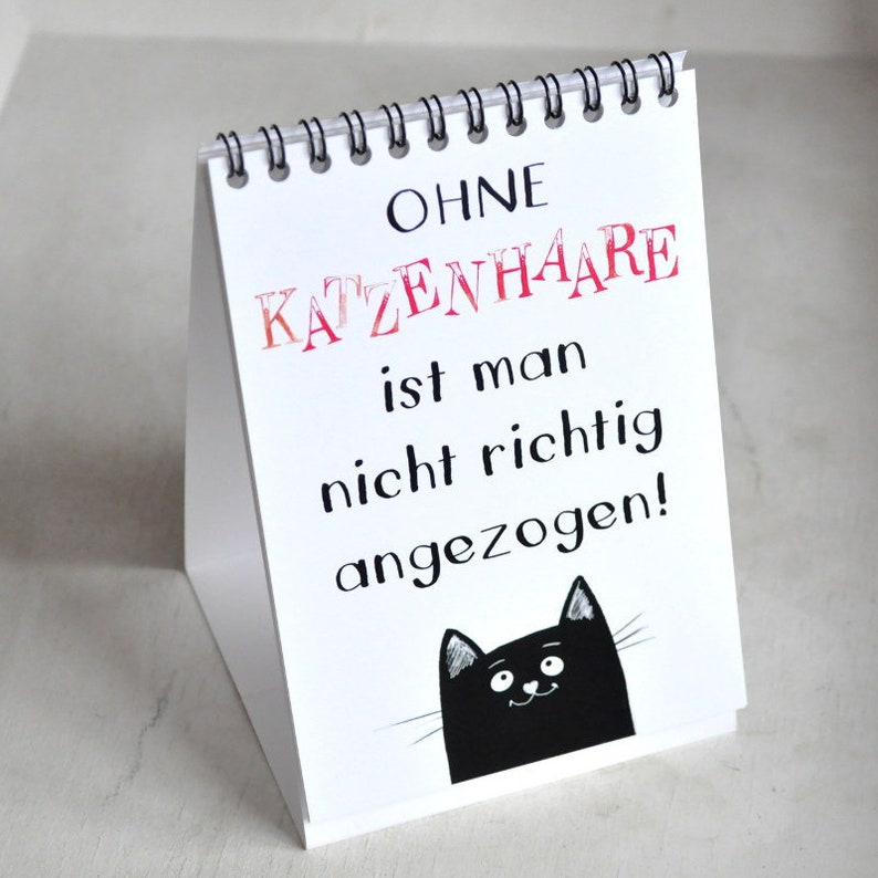 May include: White desk calendar with a black cat illustration and the text "OHNE KATZENHAARE ist man nicht richtig angezogen!"