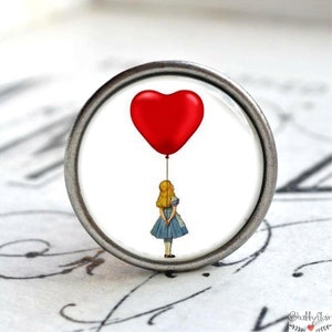 Könnte beinhalten: Ein silberner Knopf mit weißem Hintergrund, auf dem ein roter herzförmiger Ballon und ein Mädchen in einem blauen Kleid zu sehen sind, das die Schnur hält.