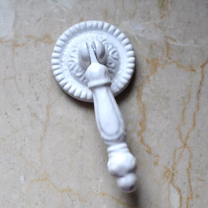 Könnte beinhalten: Weißer Keramik-Schrankknauf mit einem dekorativen, verzierten Design. Der Knauf hat eine runde Basis und einen langen, schlanken Griff.