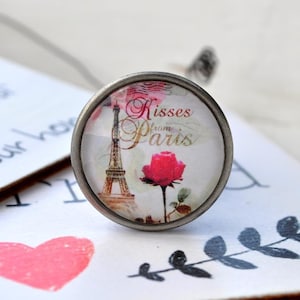 Könnte beinhalten: Ein silberner Anhänger mit einem Glascabochon, der eine Vintage-Illustration des Eiffelturms, einer rosa Rose und den Text "Kisses from Paris" zeigt.