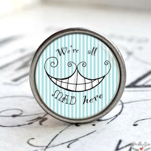 Könnte beinhalten: Ein runder, silberfarbener Knopf mit einem blau-weiß gestreiften Hintergrund. Der Knopf zeigt ein schwarzes Grinsen der Cheshire Cat mit dem Text "We're all mad here."