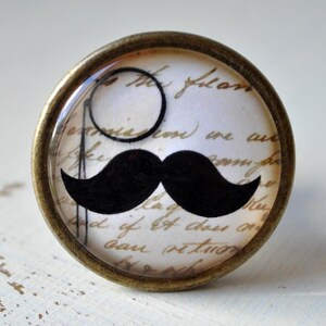 Könnte beinhalten: Ein runder, messingfarbener Knopf mit einem Glaseinsatz, der einen schwarzen Schnurrbart und ein Monokel auf weißem Hintergrund mit handschriftlichem Text zeigt.