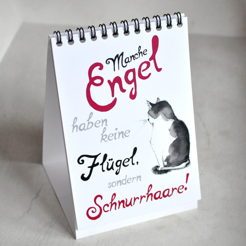 May include: A white desk calendar with a black spiral binding. The front of the calendar features a black and white illustration of a cat with the text "Manche Engel haben keine Fl&uuml;gel, sondern Schnurrhaare!" in red and black.