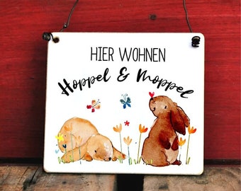 Könnte beinhalten: Quadratisches, weißes Schild mit der Aufschrift "HIER WOHNEN Hoppel & Moppel" und Illustrationen von zwei braunen Hasen, Blumen und Schmetterlingen. Das Schild hat einen schwarzen Draht zum Aufhängen.