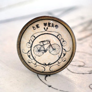 Könnte beinhalten: Ein runder, messingfarbener, dekorativer Knopf mit einem Glaseinsatz. Der Glaseinsatz zeigt eine Schwarzweißillustration eines Fahrrads mit dem Text "LE VELO" darüber.