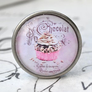 Könnte beinhalten: Ein silberner Knopf mit einem rosafarbenen und weißen Cupcake-Design. Der Cupcake hat Schokoladenfrosting und Streusel. Der Text auf dem Knopf lautet "Chocolat" und "Qualité sans Rivale".