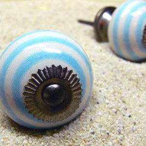 Könnte beinhalten: Zwei weiße Keramik-Schrankknöpfe mit blauen Streifen und einem silbernen Metallzentrum. Die Knöpfe befinden sich auf einer weißen Sandfläche.