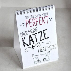 May include: White desk calendar with black text that reads "Ich bin zwar nicht perfekt, aber meine Katze liebt mich!" with a cartoon cat peeking out from the bottom of the calendar.