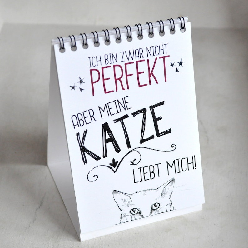 May include: White desk calendar with black text that reads "Ich bin zwar nicht perfekt, aber meine Katze liebt mich!" with a cartoon cat peeking out from the bottom of the calendar.