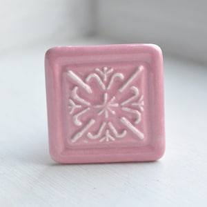 Könnte beinhalten: Ein quadratischer Keramik-Schrankknopf mit rosa Glasur und einem weißen, geprägten Blumendesign.