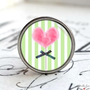 Könnte beinhalten: Ein runder, silberfarbener Knopf mit grünem und weißem gestreiftem Hintergrund. Ein rosafarbener herzförmiger Lollipop mit einem blauen Band befindet sich auf den Streifen.