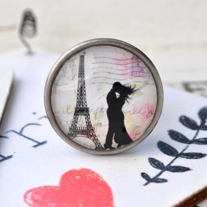 Könnte beinhalten: Ein runder silberfarbener Knopf mit einem Glaseinsatz, der eine Silhouette eines sich küssenden Paares vor dem Eiffelturm zeigt. Der Hintergrund ist eine Postkarte im Vintage-Stil mit rosa und weißen floralen Akzenten.