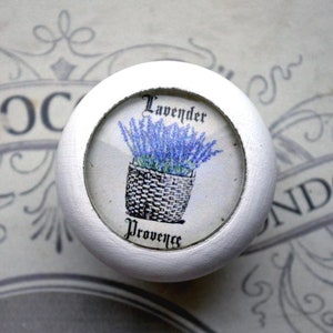 Könnte beinhalten: Runder weißer Knopf mit einem klaren Glaseinsatz, der eine Zeichnung eines Korbs mit Lavendelblüten mit dem Text "Lavender Provence" zeigt.