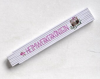 May include: White folding ruler with pink text that reads "HeimwerkerkönigIN". The ruler has a floral design with a small wooden box with a flower on it.