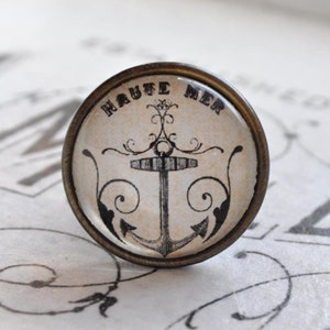 Könnte beinhalten: Runder Schubladengriff im antiken Stil mit einem nautischen Anker-Design. Der Griff hat einen cremefarbenen Hintergrund mit einem schwarzen Anker und den Worten "HAUTE MER" in kreisförmiger Anordnung. Der Metallrand hat eine Bronze-Oberfläche.