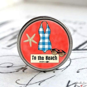 Könnte beinhalten: Ein runder, silberfarbener Knopf mit rotem Hintergrund. Der Knopf zeigt einen blau-weiß karierten Badeanzug, einen Seestern, eine Sonnenbrille und den Text "To the Beach" mit einem Pfeil, der nach rechts zeigt.