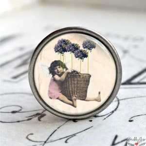 Könnte beinhalten: Ein silberner Knopf mit einem Glaseinsatz, der ein Vintage-Bild eines geflügelten Kindes zeigt, das einen Korb hält, umgeben von lila Blumen.