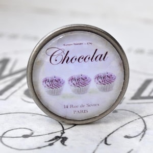 Könnte beinhalten: Ein silberner Knopf mit einem runden, weißen und violetten Design. Der Knopf zeigt drei Cupcakes mit violettem Frosting und dem Text "Chocolat", "Maison Foucher", "14 Rue de Sevres" und "PARIS".