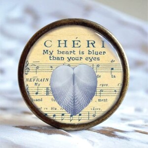 Könnte beinhalten: Ein runder, messingfarbener Knopf mit einer Glasoberfläche, die einen Hintergrund aus Vintage-Notenpapier zeigt. Der Text "CHERI My heart is bluer than your eyes" ist auf das Notenpapier gedruckt. Eine blaue herzförmige Muschel befindet sich mittig auf dem Notenpapier.
