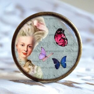 Könnte beinhalten: Runder, antik anmutender Dekorationsgegenstand mit einem Vintage-Porträt einer Frau mit gepuderter Perücke und drei Schmetterlingen. Die Schmetterlinge sind rosa, lila und blau. Der Gegenstand hat einen bronzefarbenen Metallrahmen.