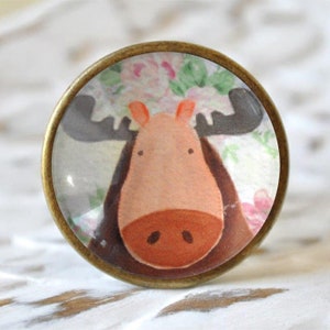 Könnte beinhalten: Ein runder, bronzefarbener Knopf mit einem aufgedruckten Cartoon-Elchgesicht. Der Elch hat braune Geweihe, eine braune Nase und ein rosafarbenes Gesicht. Der Hintergrund ist ein Blumenmuster in rosa und grün.