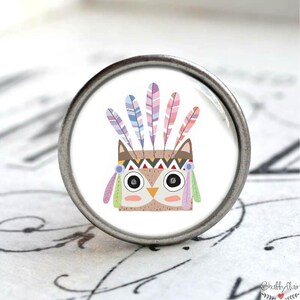 Könnte beinhalten: Ein silberner Knopf mit einem weißen Keramikeinsatz, der eine Cartoon-Eule zeigt, die einen bunten Kopfschmuck mit Federn trägt.