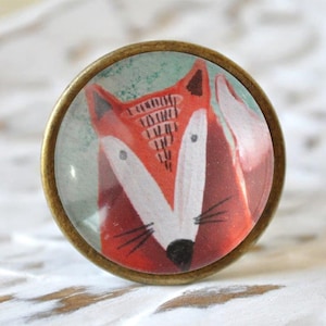 Könnte beinhalten: Ein runder, messingfarbener Knopf mit einem Glaseinsatz, der einen Comic-Fuchs mit orangefarbenem Fell, weißen Gesichtsmerkmalen und schwarzen Augen zeigt. Die Nase des Fuchses ist schwarz und er hat schwarze Schnurrhaare.