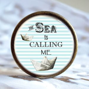Könnte beinhalten: Ein runder, bronzefarbener, dekorativer Knopf mit einem blau-weiß gestreiften Hintergrund. Der Text "THE SEA IS CALLING ME" ist in Schwarz gedruckt. Zwei Papierboote befinden sich auf dem Hintergrund.
