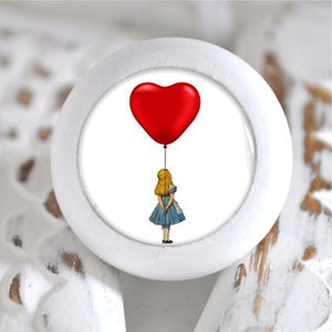 Könnte beinhalten: Weißer Keramik-Knopf mit einem roten herzförmigen Ballon und einem Mädchen in einem blauen Kleid, das die Schnur hält. Das Mädchen schaut zum Ballon auf.