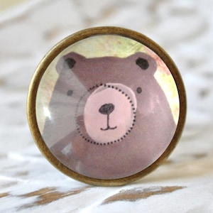 Könnte beinhalten: Ein runder, messingfarbener Knopf mit einem Glaseinsatz, der eine Cartoon-Illustration eines braunen Bären mit rosa Nase und schwarzen Augen zeigt.