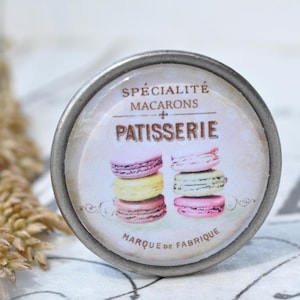 Könnte beinhalten: Ein silberner Knopf mit einem runden, weißen Etikett, auf dem "Spécialité Macarons Patisserie Marque de Fabrique" steht. Das Etikett hat einen Vintage-Look und zeigt einen Stapel bunter Macarons.