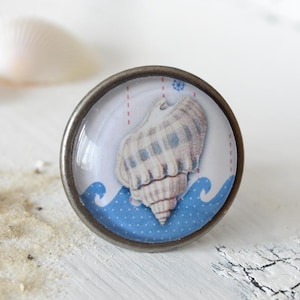 Könnte beinhalten: Ein silberner Knopf mit einem blauen und weißen Muschel-Design. Die Muschel ist weiß mit einem braunen Streifenmuster und ist von blauen Wellen mit weißen Punkten umgeben.