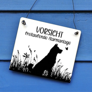 Könnte beinhalten: Ein weißes Schild mit schwarzem Text und einer Silhouette eines Hundes. Der Text lautet "Vorsicht freilaufende Alarmanlage", was "Vorsicht, frei laufende Alarmanlage" bedeutet.