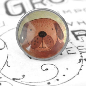 Könnte beinhalten: Ein runder Metall-Schubladengriff mit einer Aquarellillustration eines braunen Hundegesichts. Der Hund hat eine schwarze Nase, braune Ohren und einen hellgelben Hintergrund. Der Schubladengriff ist vor einem weißen Hintergrund mit schwarzer Schrift platziert.