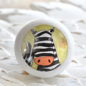 Könnte beinhalten: Weißer Holz-Knopf mit einem runden, bunten Aufkleber, der ein schwarz-weißes Zebra mit einer orangefarbenen Nase zeigt. Das Zebra befindet sich auf einem gelben Hintergrund mit einem weißen Rand.