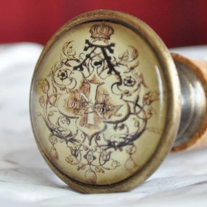 Könnte beinhalten: Ein antiker Flaschenverschluss aus Messing mit einem Glasdeckel, der mit einem detaillierten, verzierten Design verziert ist. Das Design umfasst eine Krone, einen Buchstaben 'I' und florale Motive. Der Glasdeckel ist leicht gewölbt und hat einen cremefarbenen Hintergrund.