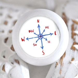 Könnte beinhalten: Weißer runder Knopf mit Kompass-Design. Der Kompass hat eine blaue Nadel und rote Beschriftung für die Richtungen: N, NO, O, SO, S, SW, W, NW.