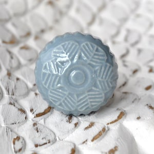 Könnte beinhalten: Ein hellblauer Keramik-Knopf mit einem erhabenen, geometrischen Design. Der Knopf hat eine leicht abgerundete Form und eine glatte, glänzende Oberfläche.