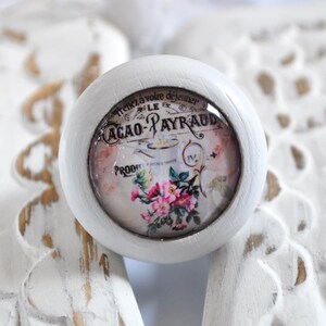 Könnte beinhalten: Weißer Keramik-Knopf mit einem runden, klaren Glaseinsatz, der ein Vintage-Design mit floralen Akzenten und dem Text "Tenez à votre déjeuner Le Cacao-Payraud" zeigt.