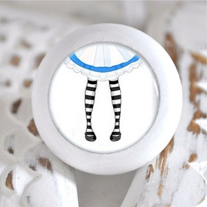 Könnte beinhalten: Weißer Keramik-Knopf mit einem runden, weißen Rahmen. Die Mitte des Knopfes zeigt ein Cartoon-Bild von einem Paar schwarz-weiß gestreiften Beinen mit einem blau-weißen Rock.