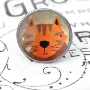 Könnte beinhalten: Eine runde, silberfarbene Anstecknadel mit einer durchsichtigen Glaskuppel, die eine Cartoon-Illustration einer orangefarbenen Tabby-Katze mit schwarzen Streifen und einer schwarzen Nase zeigt. Die Augen der Katze sind geschlossen und sie hat einen freundlichen Ausdruck.