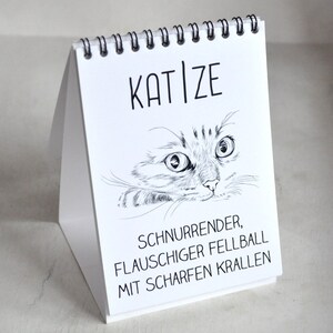 May include: A white desk calendar with a black spiral binding. The front cover features a black and white illustration of a cat's face with the text "KAT|ZE" above the cat and "SCHNURRENDER, FLAUSCHIGER FELLBALL MIT SCHARFEN KRALLEN" below the cat.