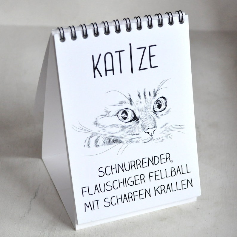 May include: A white desk calendar with a black spiral binding. The front cover features a black and white illustration of a cat's face with the text "KAT|ZE" above the cat and "SCHNURRENDER, FLAUSCHIGER FELLBALL MIT SCHARFEN KRALLEN" below the cat.