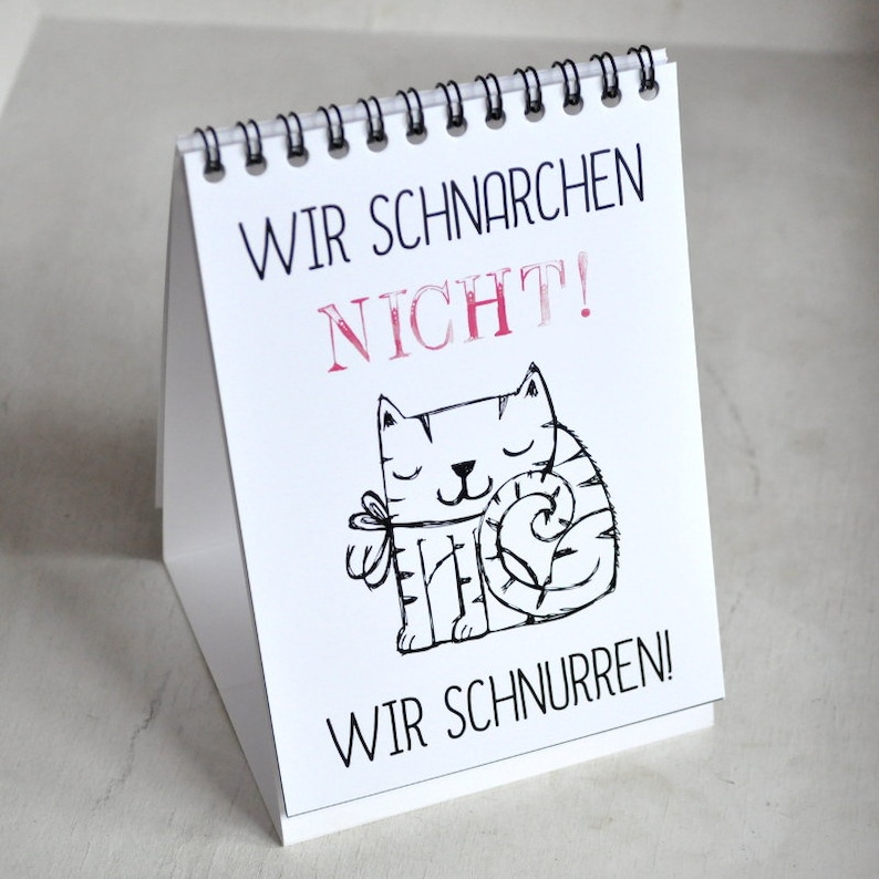 May include: A white desk calendar with a black spiral binding. The calendar features a black and white illustration of a cat with the text "Wir schnarchen nicht! Wir schnurren!" in German.