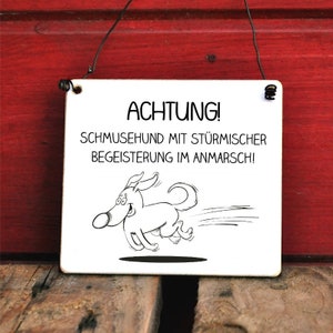 Könnte beinhalten: Ein weißes Schild mit schwarzem Text und einem laufenden Comic-Hund. Der Text lautet "ACHTUNG! SCHMUSEHUND MIT STÜRMISCHER BEGEISTERUNG IM ANMARSCH!"
