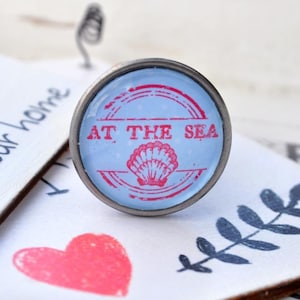 Könnte beinhalten: Ein silberfarbener Metallknopf mit einem blauen und roten Design. Der Knopf zeigt eine Muschel und den Text "AT THE SEA".