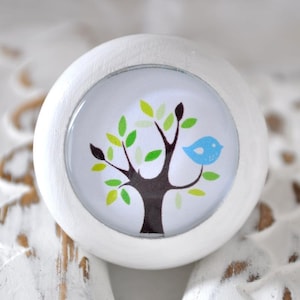 Könnte beinhalten: Runder weißer Knopf mit einem braunen Baum und einem blauen Vogel auf weißem Hintergrund. Der Knopf ist ein dekorativer Akzent für einen Schrank oder eine Schublade.