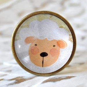 Könnte beinhalten: Ein runder, messingfarbener Knopf mit einem Glaseinsatz, der eine Cartoon-Illustration eines weißen Schafs mit rosa Nase und Rouge zeigt.