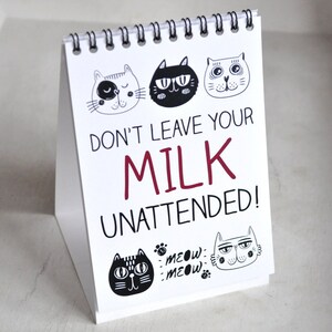 May include: White sign with black and white cat illustrations and the text "Don't leave your milk unattended!" in red and black text. The sign has a spiral binding and is standing upright.
